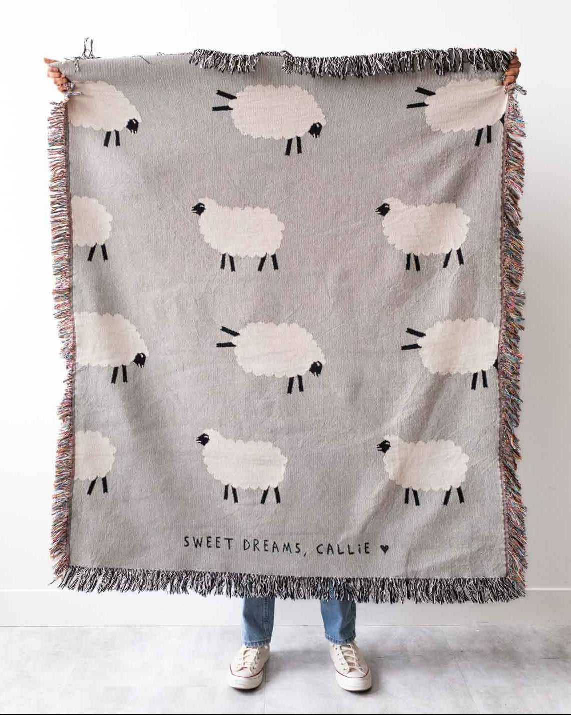 A light grey cotton blanket with a pattern of frolicking sheep, personalized with the text 'SWEET DREAMS, CAITIE' at the bottom.
