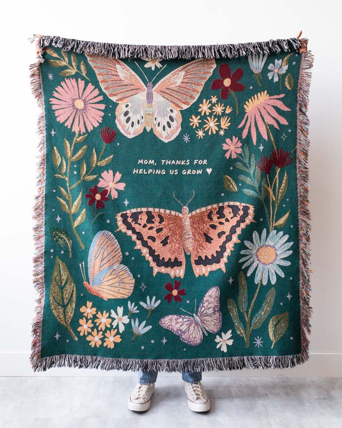A teal green cotton blanket with a floral and butterfly pattern, featuring a personalized message, displayed by a person standing against a white background.