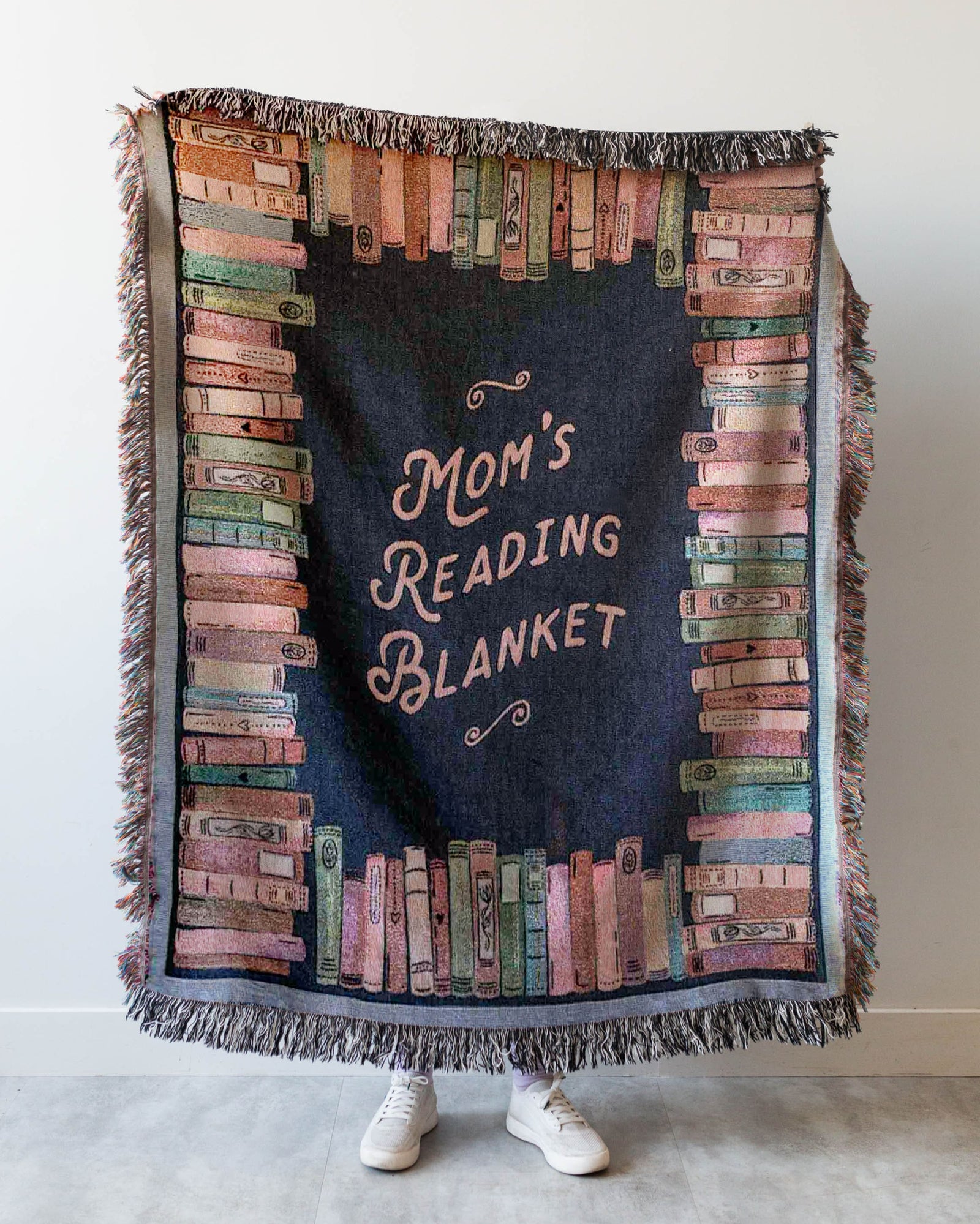 Blanket with 'Mom's Reading Blanket' text and book design, held by a person wearing white shoes.