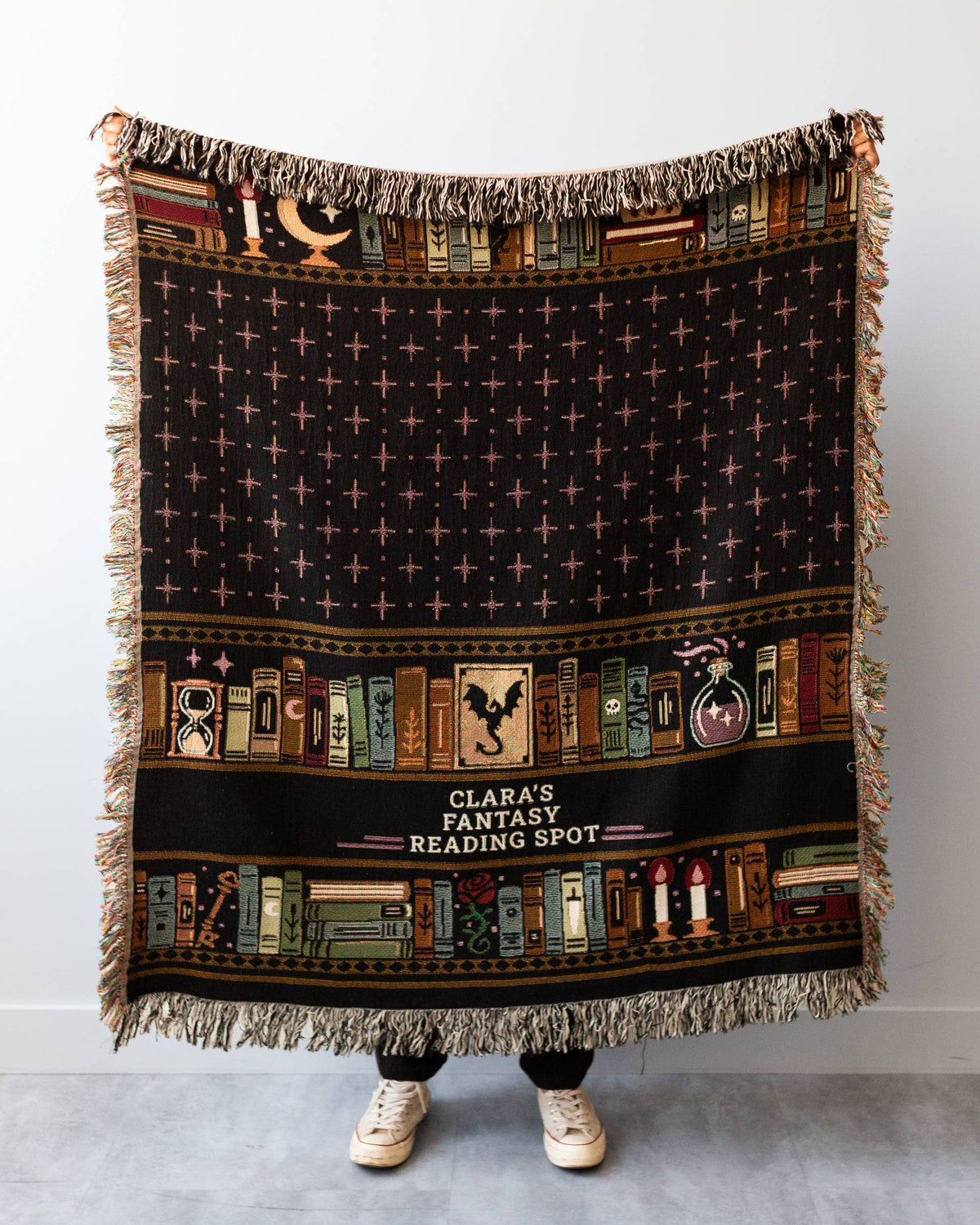 A person holds up a large woven throw blanket featuring a dark fantasy library theme with the text 'CLARA'S FANTASY READING SPOT'. The blanket has a black background with a grid of stars, a horizontal band of colorful book spines, and fringed edges.