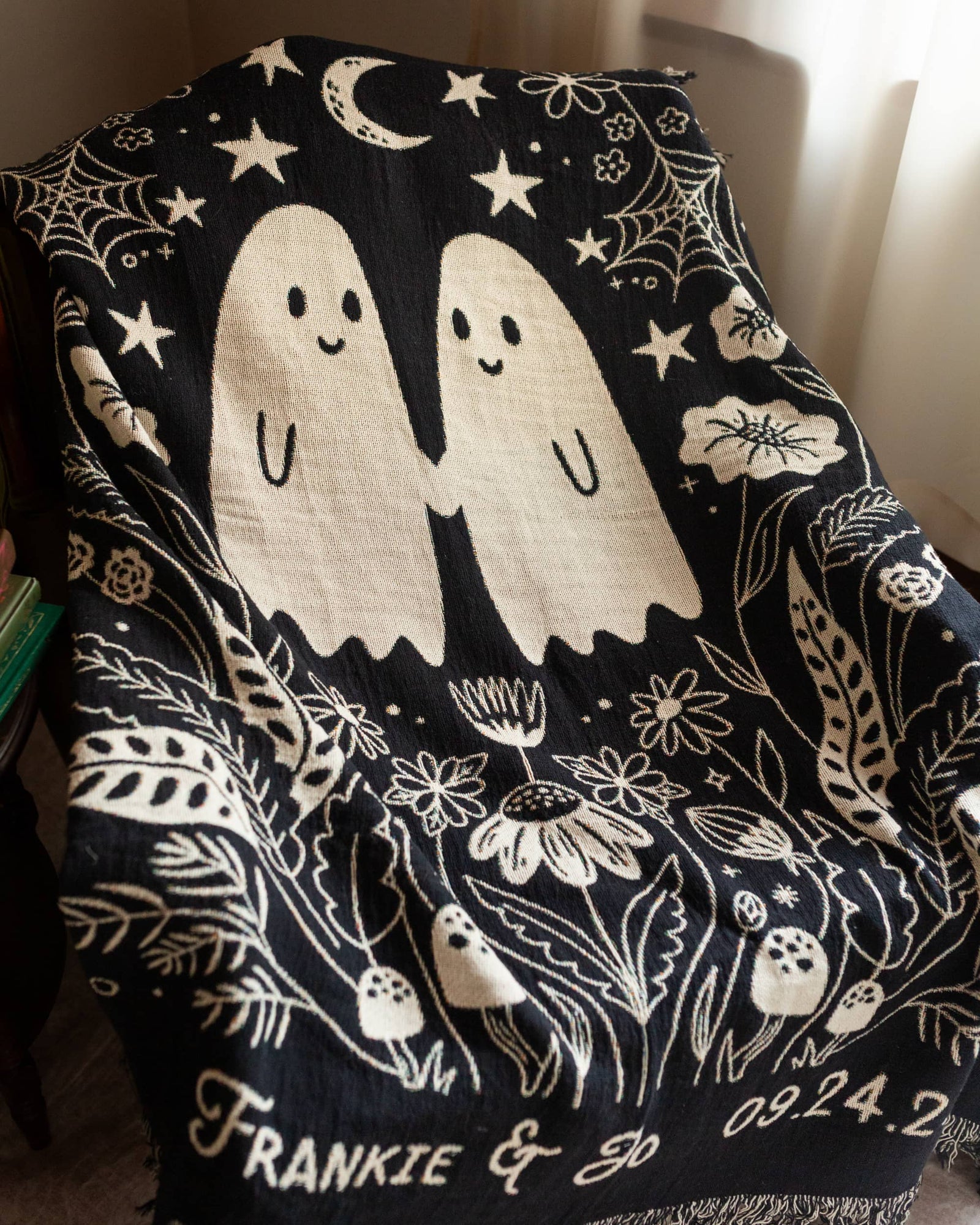 Black woven throw blanket with white ghost design and personalized names text, held by a person wearing white sneakers.