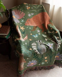 Person holding a decorative woven throw blanket with woodland animal and floral patterns on a white background
