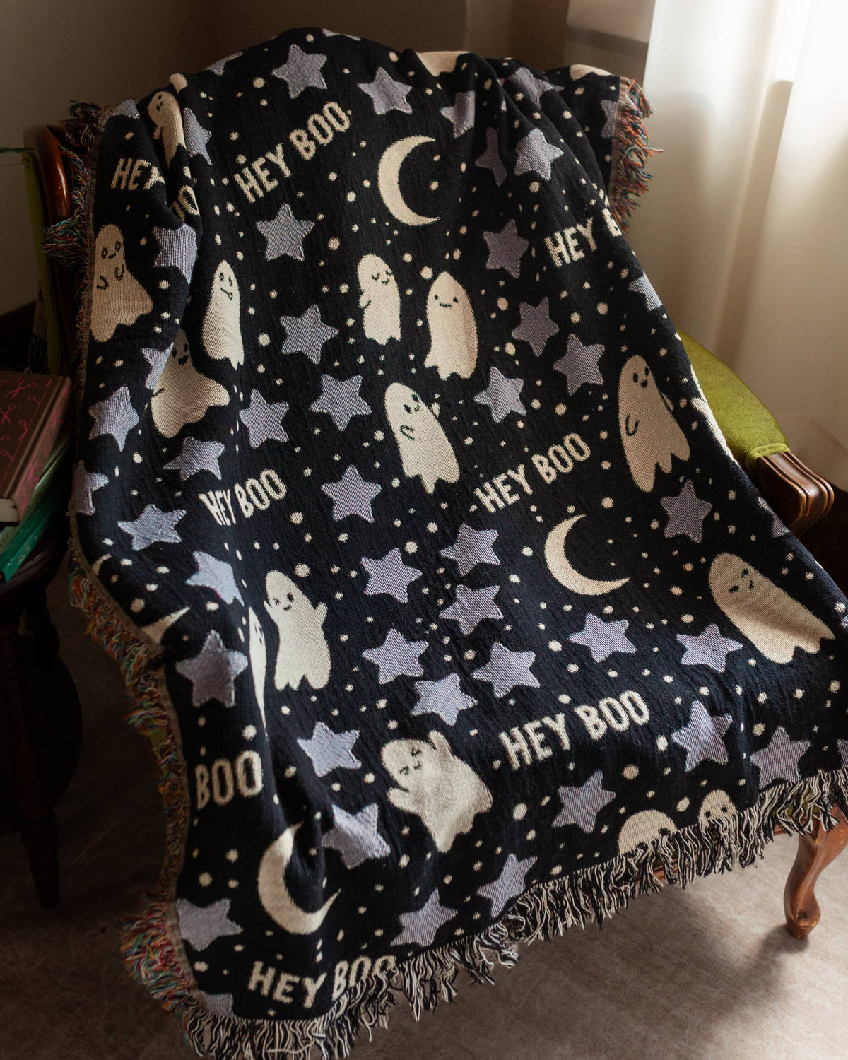 A black woven throw blanket with a Halloween pattern featuring white and gray ghosts, stars, crescent moons, and the words 'HEY BOO', draped over a chair in an indoor room.