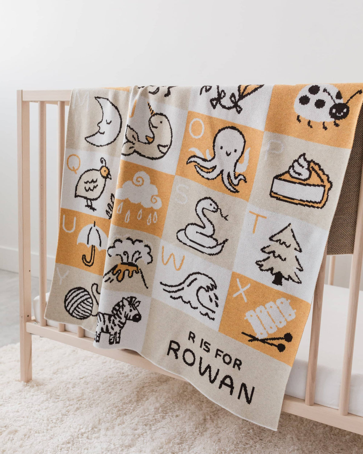 Baby blanket with alphabet patterns and 'R is for Rowan' text draped over a crib.