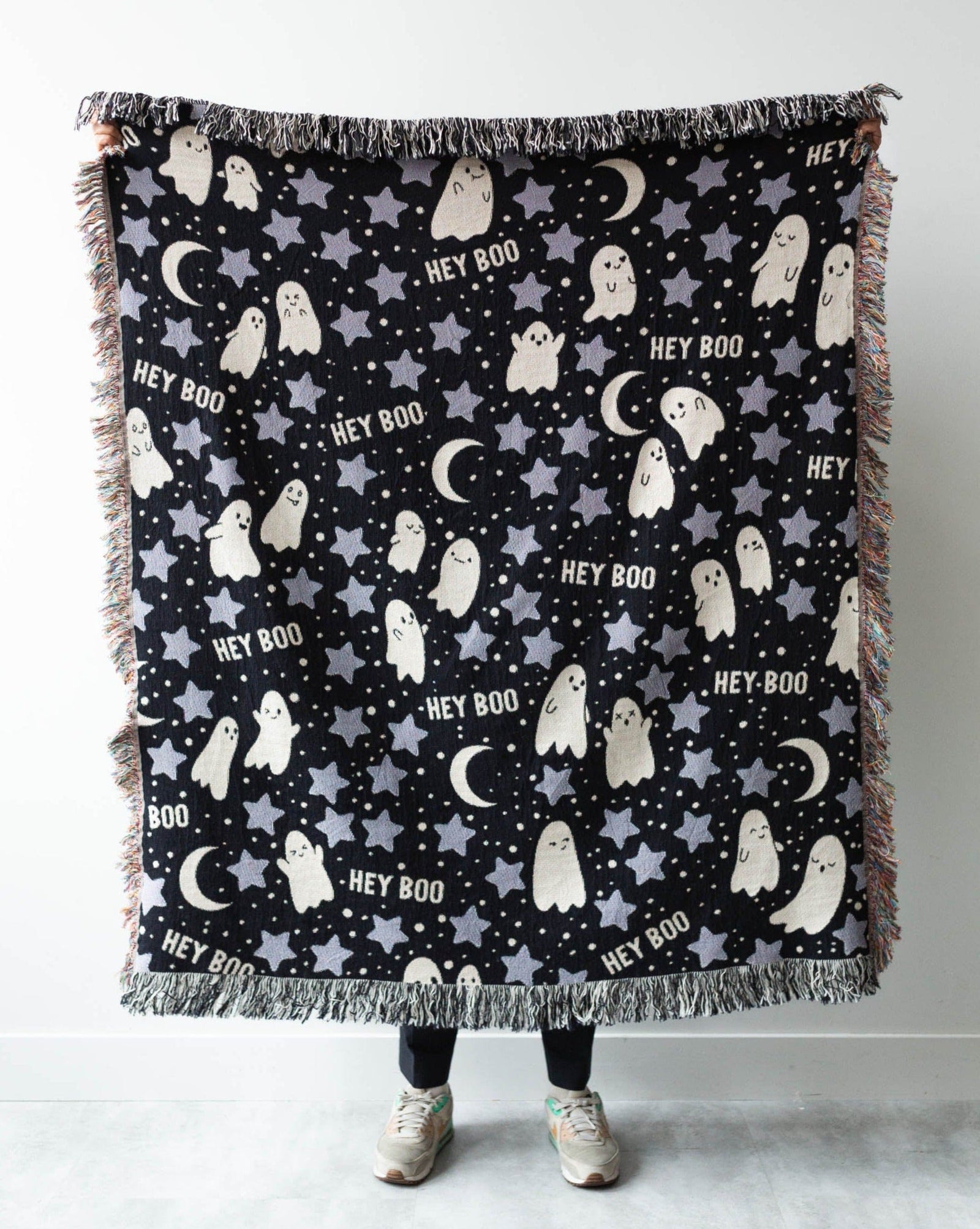 A black woven throw blanket with a Halloween pattern featuring white and gray ghosts, stars, crescent moons, and the words 'HEY BOO', draped over a chair in an indoor room.